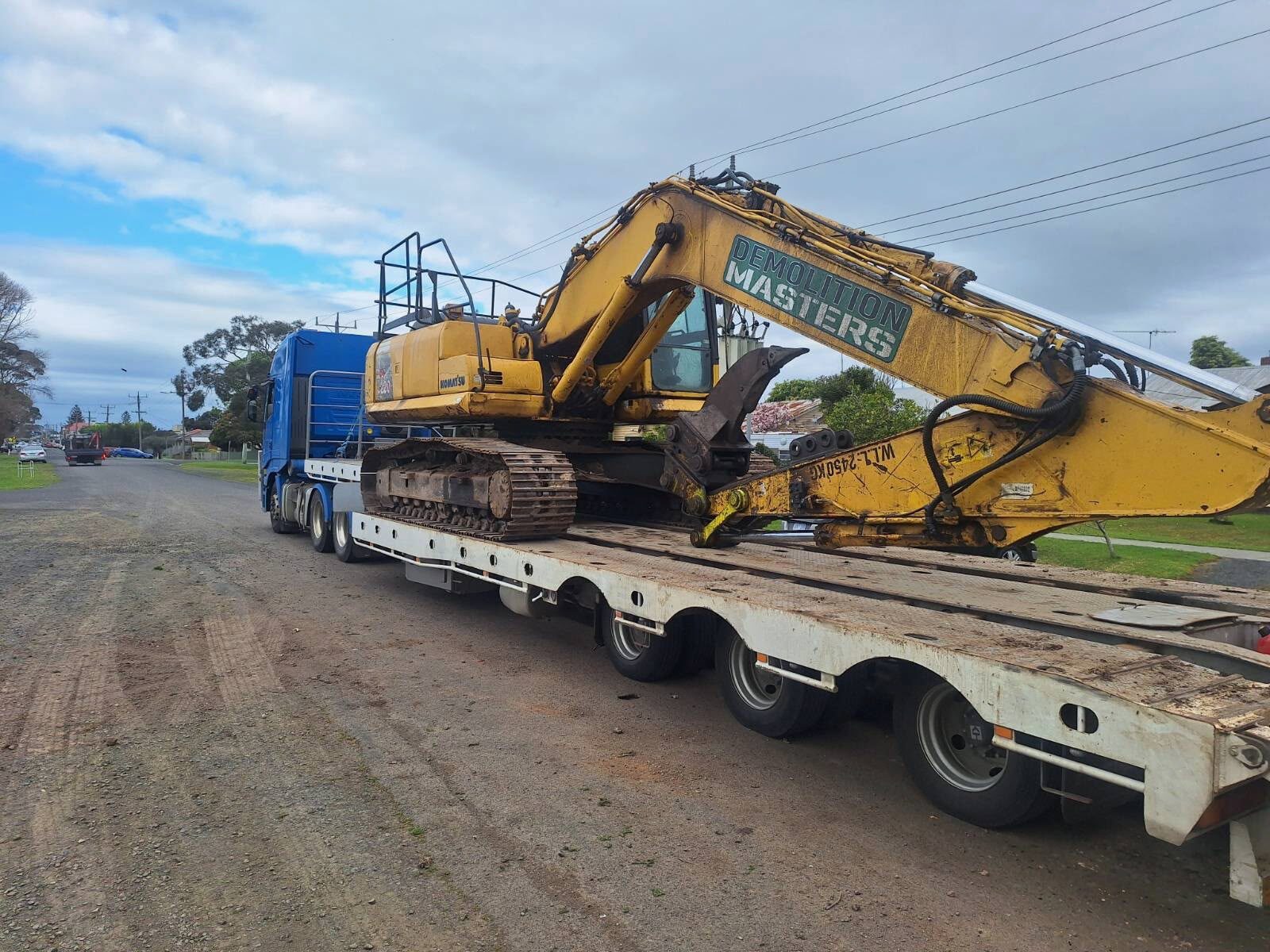 Yellow Demolition Masters Komatsu excavator on a float trailer on a gravel road
